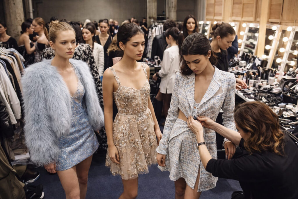 Models wearing designer outfits backstage at fashion week while stylists adjust clothing before runway